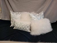Five white decorative accent pillows including two with silver abstract floral patterns, one rectangular pillow with silver geometric designs, one fluffy fur pillow, and one feathered pillow. Pillow set displayed on black and beige backgrounds.