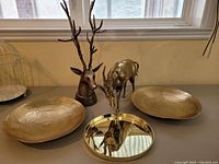 Photo showing two brass deer head sculptures with one having large antlers, brass antelope figurine, and three brass decorative dishes on table near window.