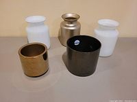Five decorative vases grouped on tabletop: two white opaque glass vases, one metallic gold vase, one dark brown ceramic vase, and one more white opaque glass vase.