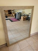 Frontal photo of large rectangular mirror with brushed gold frame reflecting a tiled room with furniture and boxes.