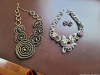 Two necklaces and a pair of earrings displayed on a brown surface. Left necklace is bronze faux heirloom with spiral rings, right necklace is rhinestone flower design with matching earrings above it.