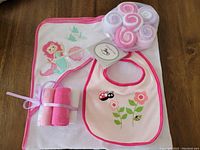 Photo showing hooded towel with pink trim and mermaid embroidery, bib with flower and ladybug embroidery, and six rolled face cloths in pink and white tones all together on a wooden surface.