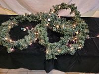 Six artificial evergreen garlands arranged on a black cloth surface, showing green foliage and warm white string lights turned on.