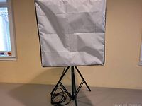 Square softbox light reflector setup on tripod stand in a room with beige walls and window showing some natural light.