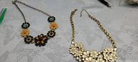 Both gold-toned metal necklaces shown side by side on patterned white surface, one with multicolored floral stones and the other with white enamel flowers.