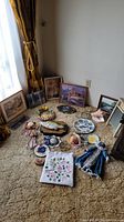 Wide shot showing collection spread on carpet near window with curtain and baseboard vent; includes framed pictures, dolls, plates, embroidery, and a flower wreath