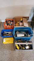 Six containers of assorted tools on carpet including plastic bins, metal toolbox, and yellow toolbox. Various hand and power tools are seen inside containers.