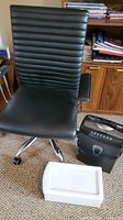 Black ribbed swivel office chair, paper shredder, and white storage bin shown from front with office supplies in background.