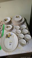 Photo showing the full set of Ridgway ironstone china dishes with yellow and blue rose pattern, including stacked dinner plates, smaller plates, six bowls, and six cups, all with gold trim.