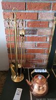 Full brass fireplace tool set with stand and copper kettle shown with brick fireplace background.