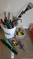 Photo of a white RONA bucket filled with assorted gardening tools including hand rakes, pruning shears, and other small tools beside a clear plastic container with gardening accessories on carpeted floor.