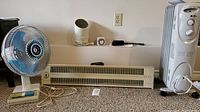 Photo showing a medium oscillating fan with blue/white grille, a small white fan, a standard 1000 watt floor heater, and two mechanical timers on a shelf.