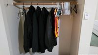Image showing five men's coats and jackets in black and a high-visibility safety vest hung on a wooden rod with empty white and black coat hangers on the right side.