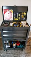 Black wheeled tool chest with open top showing screwdrivers, tape rolls, and a Diablo circular saw blade package inside.