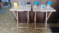 Two wooden foldable TV trays each with multiple cartoon character mugs and cups on top, background kitchen counter.