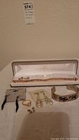 Jewelry items arranged on surface including chain necklace in box, multiple pairs of earrings, wide cuff bracelet, and rings.