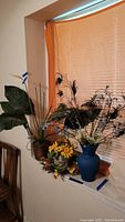 Wide view of three faux flower arrangements placed on window sill, including blue vase with peacock feathers, brown pot, and basket arrangement.