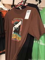 Brown Eric Clapton concert t-shirt hung on pink hanger in front of other t-shirts on black hanger.