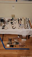 Wide view of the costume jewelry lot laid out on table and floor showing variety of necklaces, bracelets, earrings, watches, and miscellaneous items.