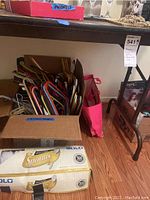 Photo shows a box filled with colorful plastic and wire hangers, an opened box of 500 Solo disposable spoons, and partially visible Victoria's Secret bags and boxes near the floor under a shelf.