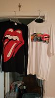 Black long sleeve Rolling Stones t-shirt with large red and white tongue and lips logo on front, hanging next to white short sleeve Rolling Stones t-shirt with colorful lips graphic