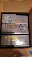 Three unopened tin boxes of Double Twelve Mexican Train Dominoes inside a cardboard box, stacked together with visible printed text and images on tins.