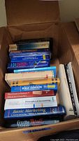 Photo showing a cardboard box filled with various academic textbooks on topics including marketing, accounting, human resources, and economics, stacked horizontally and vertically.