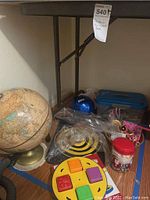 Photo showing the globe on a stand, the yellow educational toy with colored letter blocks, a blue plastic dog toy, and a blue box (likely art supplies) on the floor under a table.