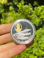 Front side of coin showing vintage biplane and gold-colored inset portrait, held between fingers with trees in background.