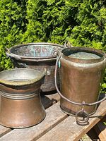 Three antique Arts & Crafts era hand hammered copper pots displayed on wooden surface outdoors against greenery, showing overall condition and patina.