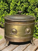Front view of oval brass antique coal box showing lid with finial, decorative horizontal bead bands, medallion accents, and claw feet, placed outdoors.