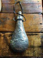 Front view of antique pewter powder flask showing wildlife scene with deer and stag in embossed design and fabric strap.