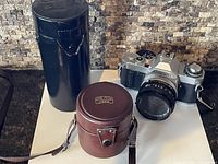 Canon AV-1 camera with leather strap, black cylindrical case for a lens, and separate brown Carl Zeiss Jena lens case with label.