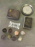 Photo showing metal match case, round metal tray, metal match safe with portrait, vintage British Columbia chauffeur license plate, and assorted metal pins.