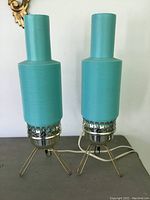 Front view of both turquoise plastic rocket-style lamps on metal tripod stands on table surface.