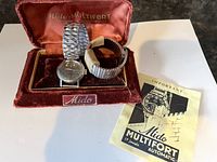 Photo of the open red velvet Mido Multifort branded case with the watch and two metal expandable bands inside, alongside the instruction booklet.