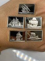 Photo of six rectangular silver bars with detailed ship engravings on black backgrounds, displayed on hand for size reference.