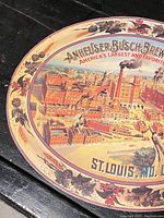 Close-up of upper left portion of the oval Anheuser Busch cardboard sign showing detailed brewery illustration and grapevine border.