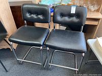 Front view of two black barstools with leather-like upholstery showing seating area and button tufting on backs, chrome tubular bases on carpeted floor.