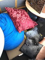 Photo of the bean bag chair draped with four different pillows: blue bean bag with suede-like fabric, red floral decorative pillow, beige pillow, and black pillow with palm leaf design.