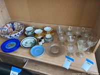 Wide view of the shelf with mixed Asian ceramic bowls, tea cups, saucers, and blue Fiesta ware saucer along with clear glassware plates and etched glasses.