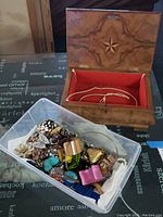 Full lot showing a wooden jewelry box with red felt interior and a plastic container filled with assorted costume jewelry items including brooches, rings, pins, and more.