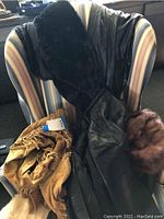 Black leather coat, black leather pants, tan suede coat with tag, brown fur hat, and black fur garment on striped chair