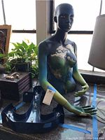 Painted mannequin sitting near window with a nature-inspired design on its torso and blue-green gradient arms. Lighted anchor wall decoration placed in front of mannequin's lap.