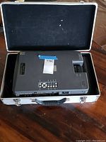 Image showing top view of Sharp XG-NV1U liquid crystal projector inside a silver hard case with foam lining. The projector has various control buttons and a visible lens area.