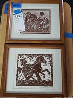 Two framed artwork prints displayed together on a wooden surface, showing front view of artwork and frames.