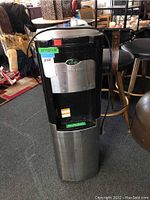 Full view of upright black and stainless steel Viva water dispenser in a room with furniture and other items.