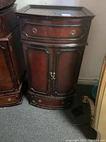 Full view of a wooden jewelry cabinet showing a dark wood finish with a curved front, a top drawer with two metal ring handles, and a lower double-door cabinet with metal knobs.
