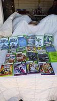 Photo showing all 20 Xbox 360 game cases arranged on a white bedspread, titles visible including Rock Band 2, NBA 2K11, Grand Theft Auto IV, and others.