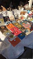 Overview photo of 45 RPM vinyl singles laid out on a white cloth surface, showing multiple records with various colorful sleeves and labels.